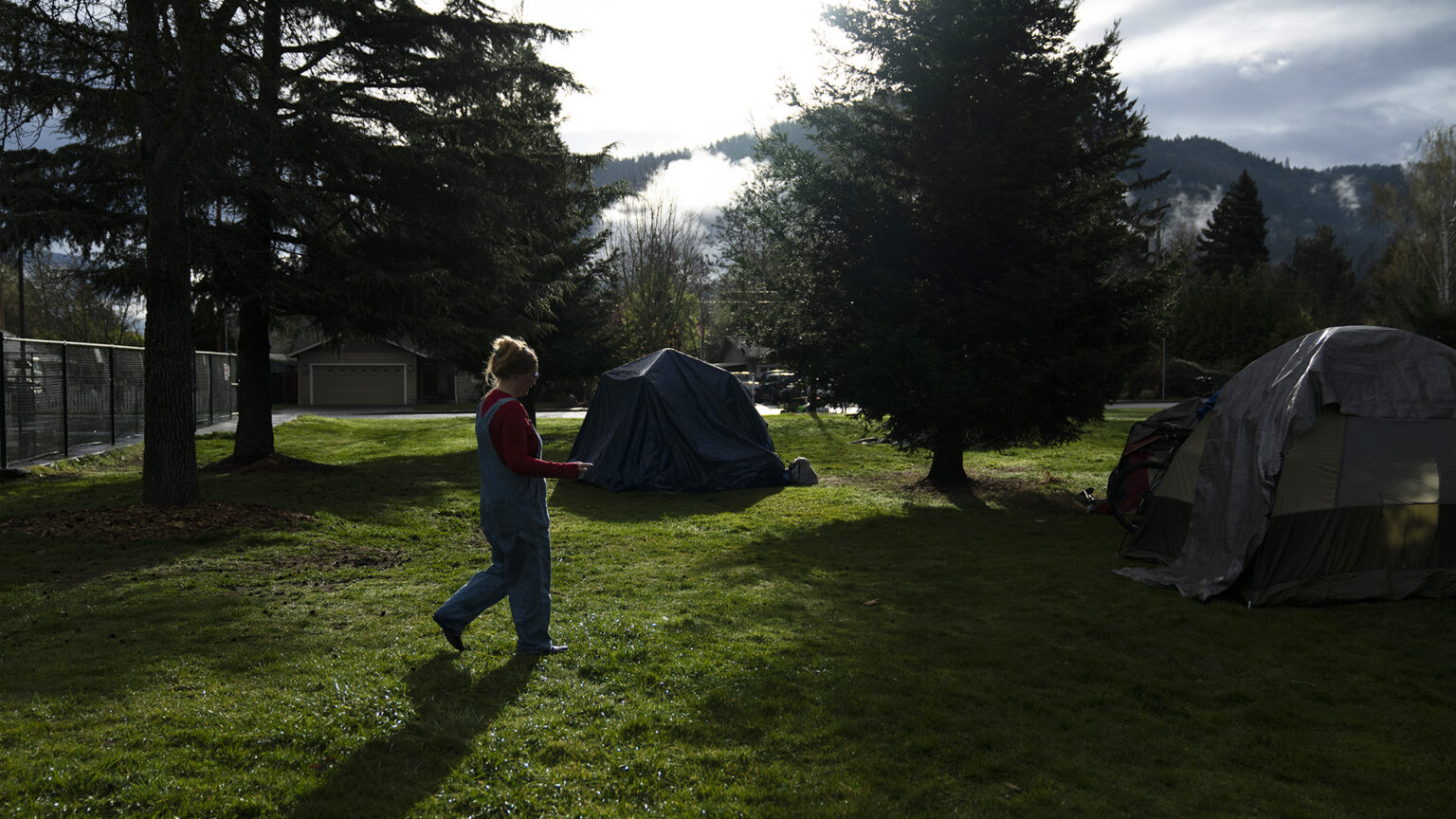 Cassy Leach walks along a lawn with multiple domed camping tents pitched among several trees, with a fence, garage and trees in the background and sun shining through low clouds along a tree-covered range.
