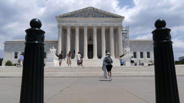 People stand on a steps and a plaza outside the west façade of the U.S. Supreme Court Building with marble masonry, a portico of columns flanked at their base by two sculptures of seated figures, and a pediment with a of sculptures of figures, framed by two metal parking bollards in the foreground.