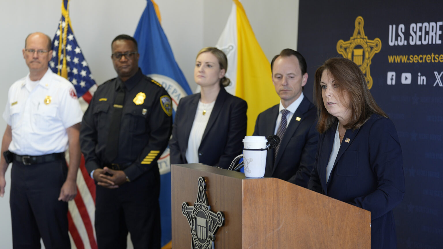 Kimberly Cheatle speaks while standing behind a wood podium with the seal of the U.S. Secret Service affixed to its front, with four other people standing to her side in a room with the U.S., Wisconsin and Milwaukee flags in the background.