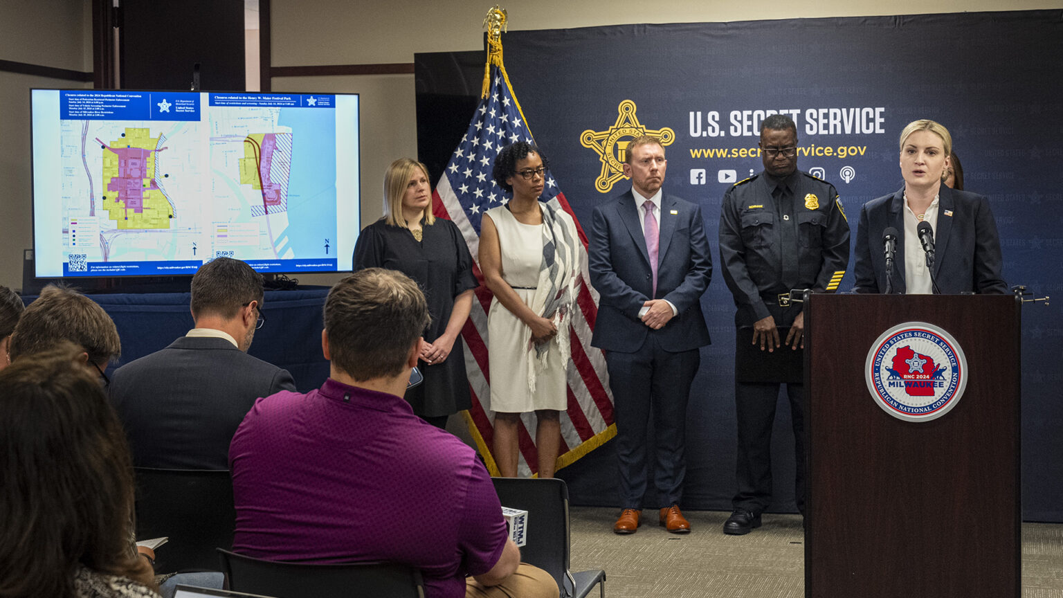 Audrey Gibson-Cicchino stands and speaks into two microphones attached to a wood podium affixed with a logo of the 2024 Republican National Convention, with other people standing behind her in front of a U.S. flag and a photo backdrop with a graphic of the badge of the U.S. Secret Service and next to a video monitor showing multiple maps, faced by people seated in stackable chairs in the foreground.