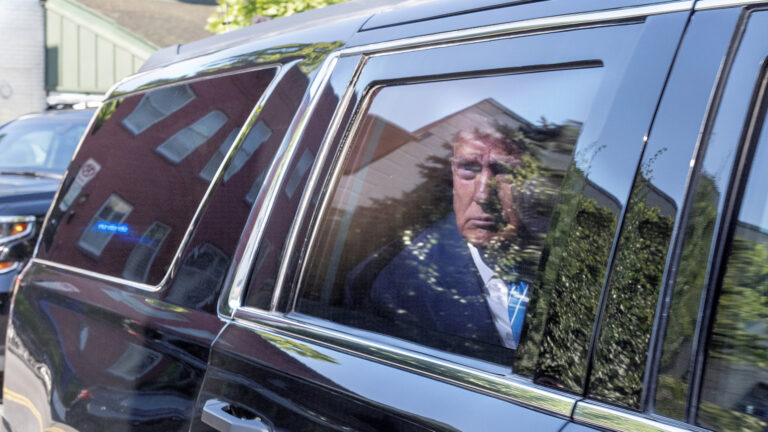 Donald Trump looks out a rear passenger-side window while seated in a SUV limousine, with tree leaves and buildings reflecting off the paint and glass surfaces of the vehicle.