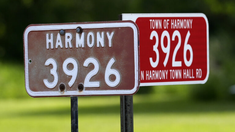 Metal address markers that read Harmony 3926 and Town of Harmony 3926 N Harmony Town Hall Rd are affixed to the top of metal rods, with an out-of-focus lawn and foliage in the background.