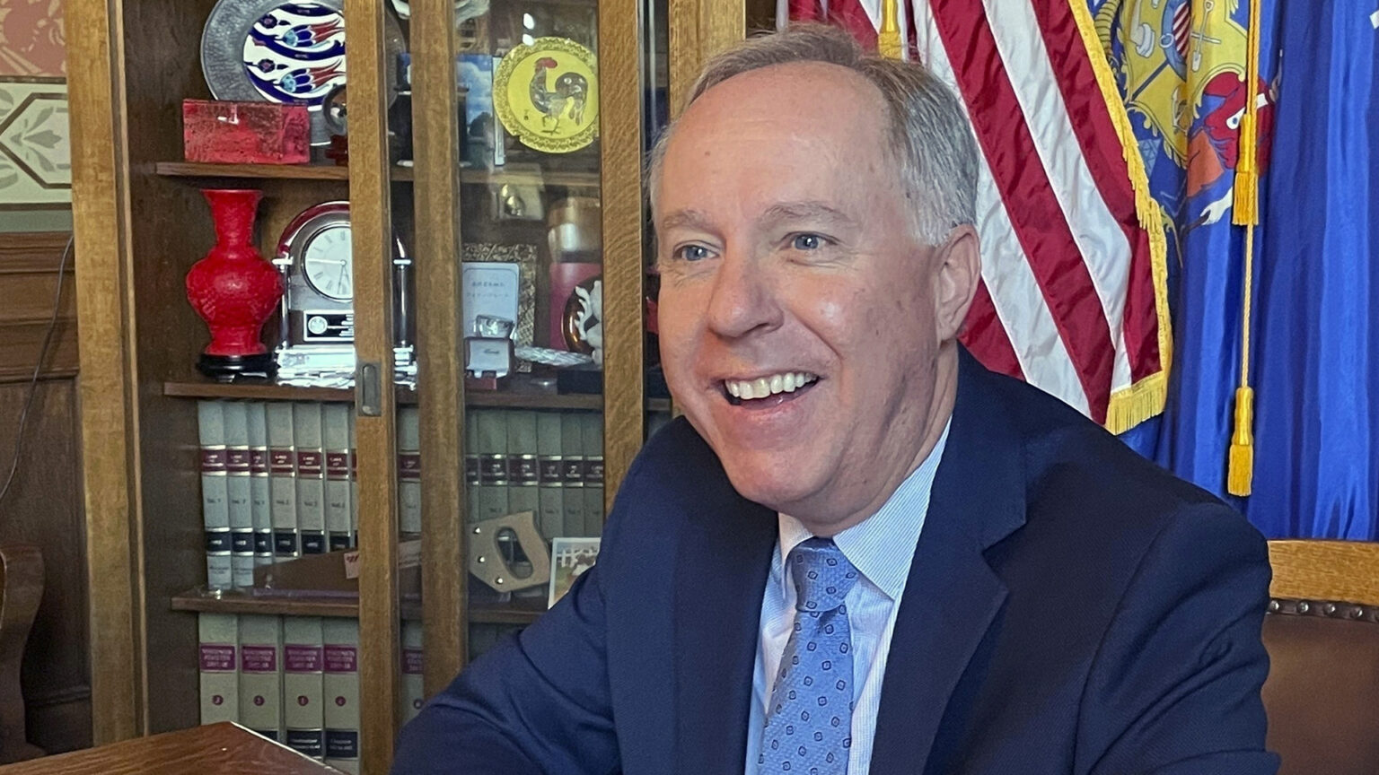 Robin Vos sits in a wood and leather curved back chair inside a room with the U.S. and Wisconsin flags, and a cabinet with law books and decorative items.