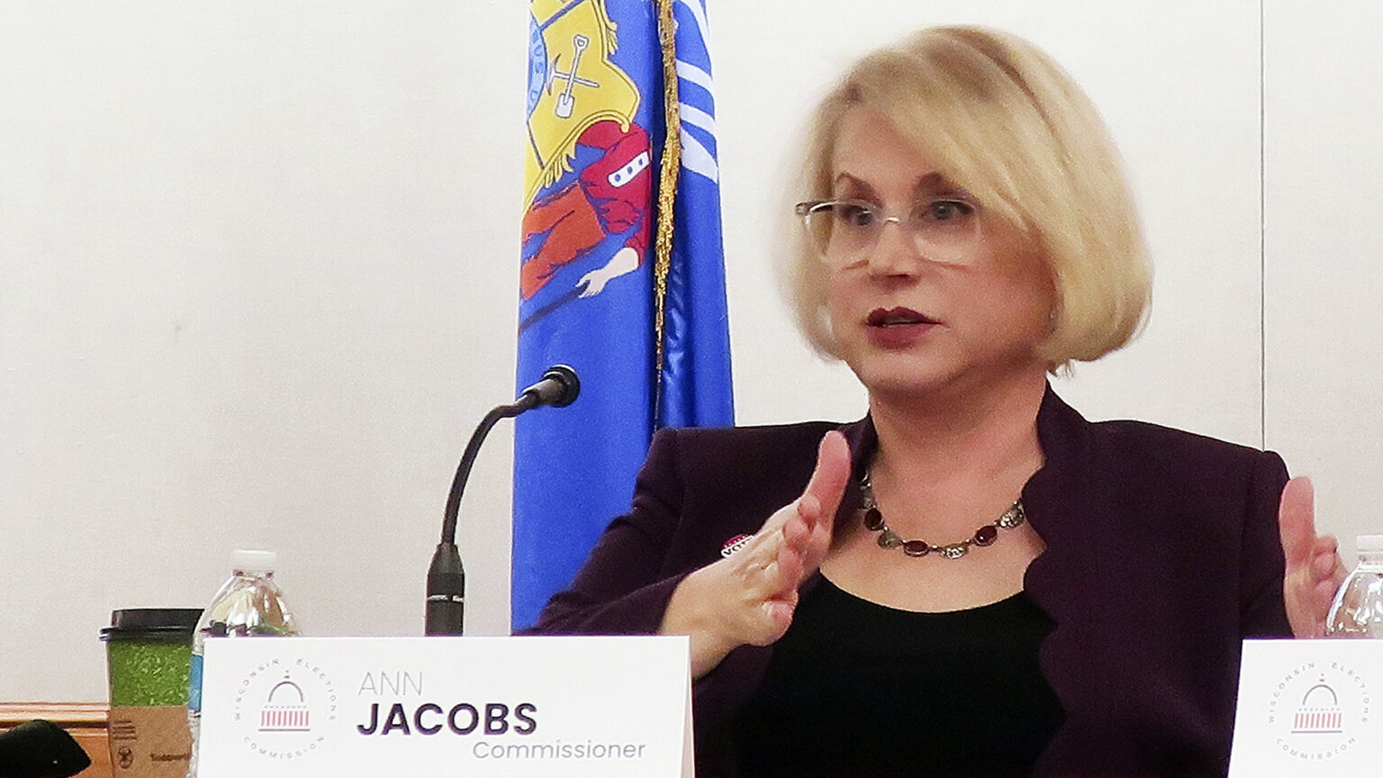 Ann Jacobs gestures with both hands and speaks into a microphone while seated at a table with a folded paper sign showing the logo of the Wisconsin Elections Commission and the words Ann Jacobs and Commissioner on its surface alongside a disposable coffee cup and plastic water bottle, with a Wisconsin flag in the background.