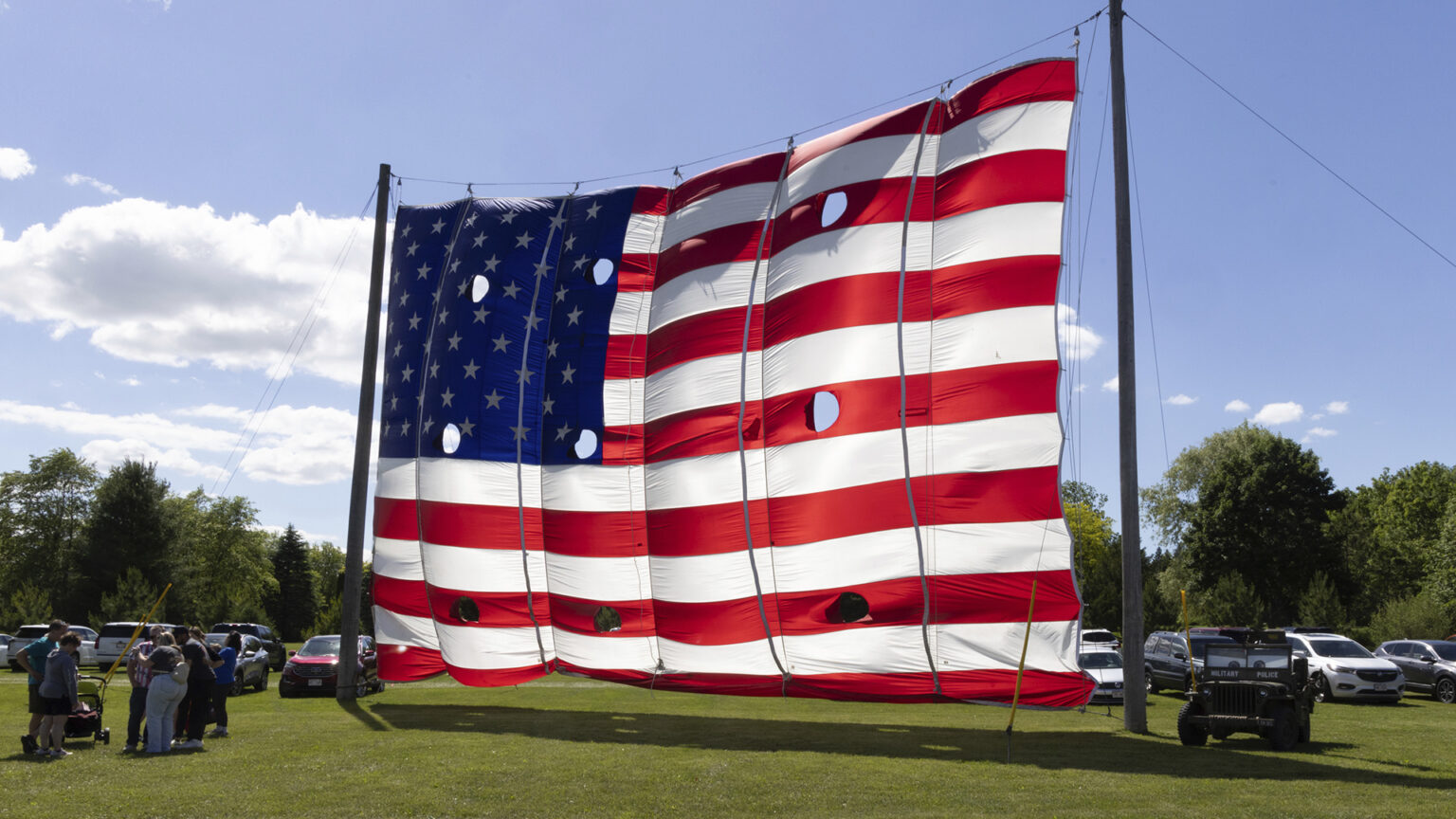 A multi-story tall U.S. flag with nine circular holds in it to allow for wind passage is mounted on a system of ropes and pulleys on two tall wood poles, with several people standing near the base of one and a UTV parked near the base of another, with parked cars and trees in the background.