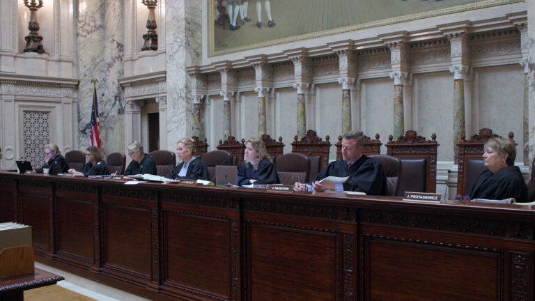 From left to right, Jill Karofsky, Rebecca Dallet, Ann Walsh Bradley Annette Ziegler, Rebecca Bradley, Brian Hagedorn and Janet Protasiewicz sit at a judicial dais in a row of high-backed leather chairs behind them, with another row of high-backed wood and leather chairs behind them, in a room with marble masonry, a U.S. flag and a large painting.