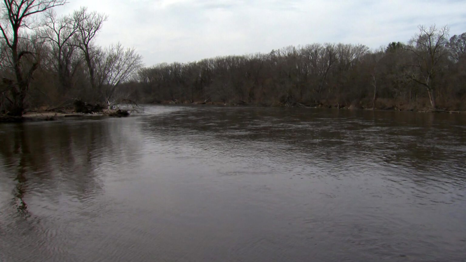 Leafless deciduous trees line both shores of a river under a cloudy sky.