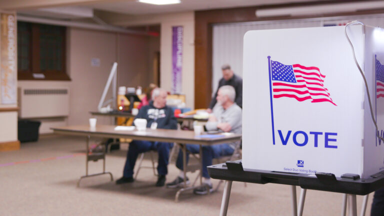 An illustration of the U.S. flag and the word Vote are printed on collapsible privacy barriers of a voting booth inside a polling place, with out-of-focus people seated at a folding table in the background.