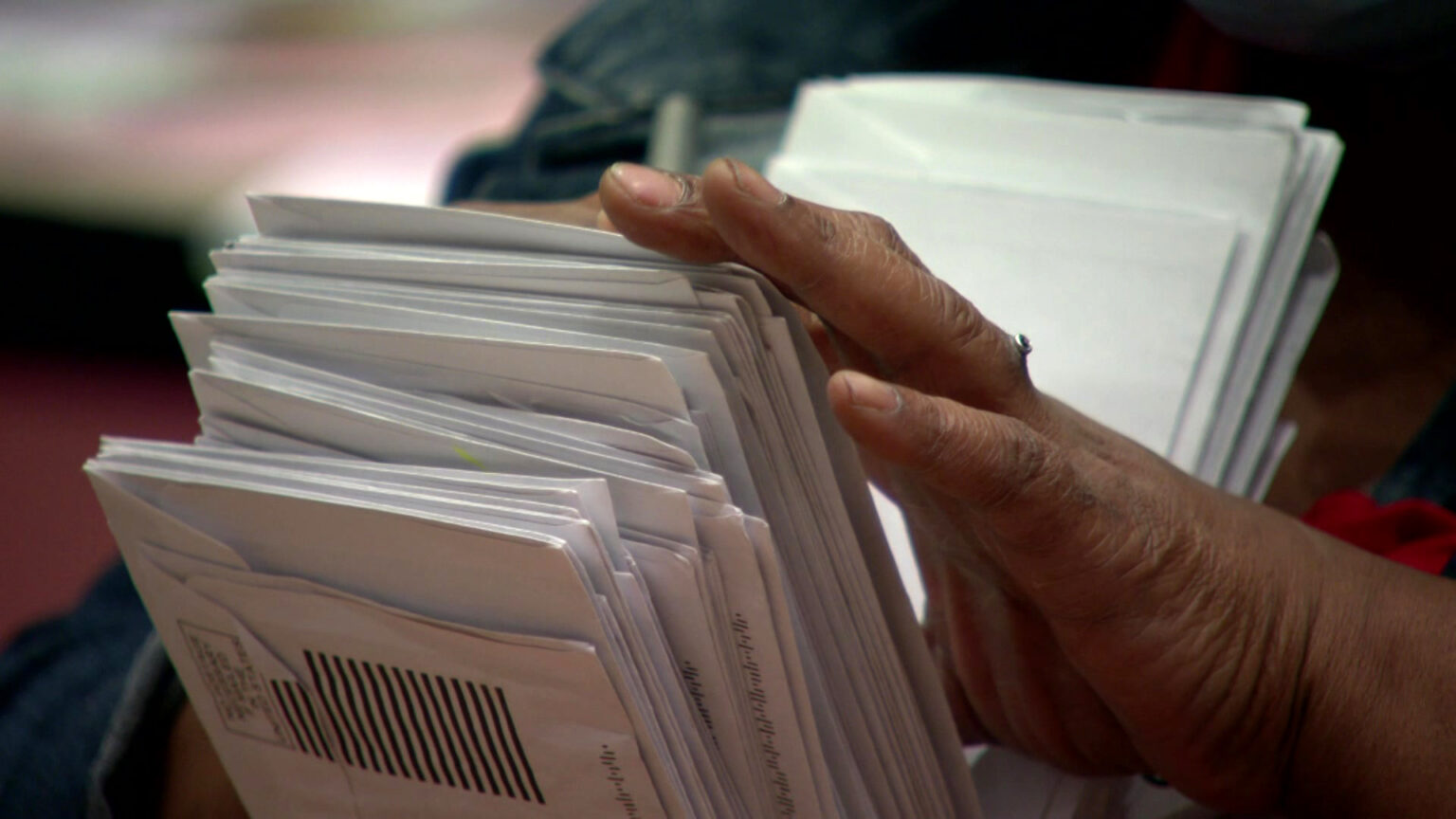 A hand touches the edge of an absentee ballot envelope in a stack held on a lap.