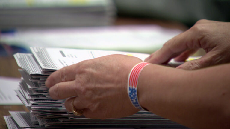 Two hands grasp an absentee ballot envelope from the top of a stack of ballot envelopes on a table.