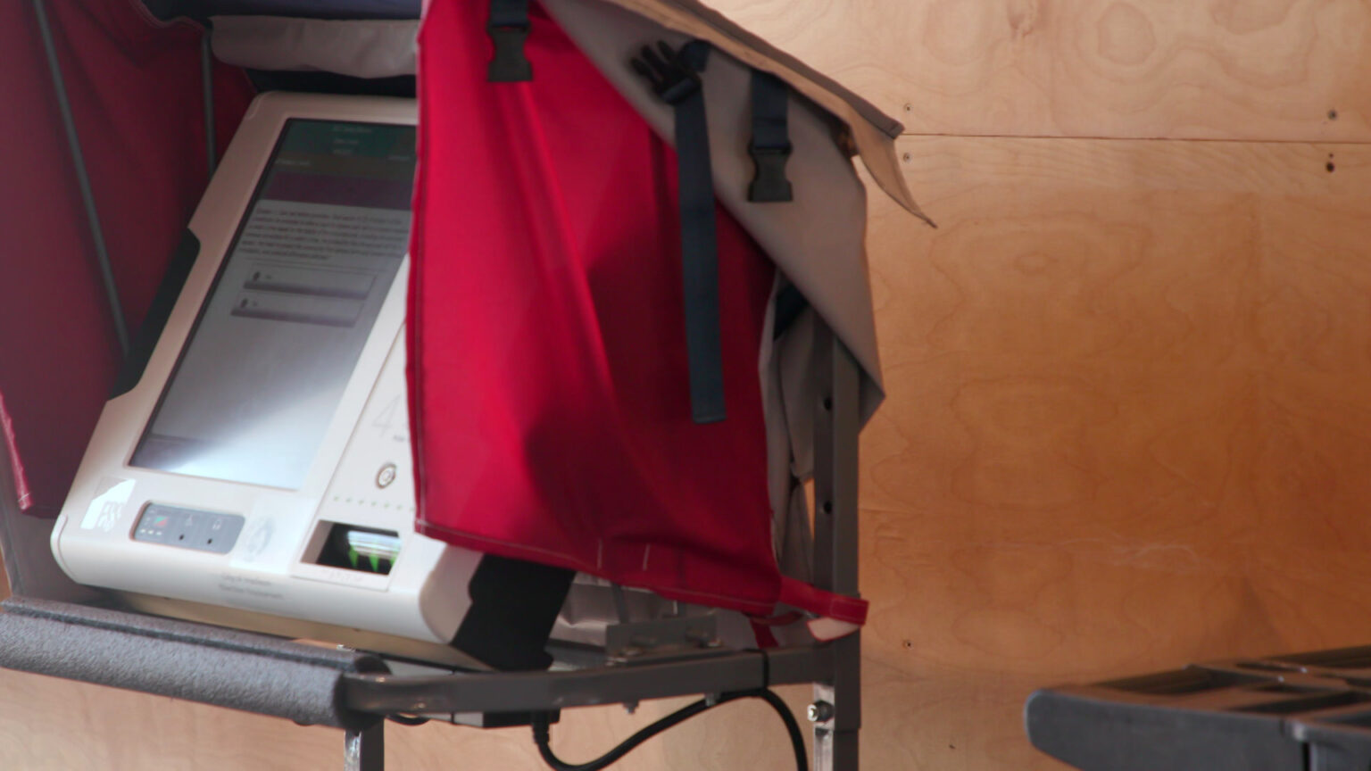 A touchscreen printed ballot voting machines stands in a room with wood-paneled walls.