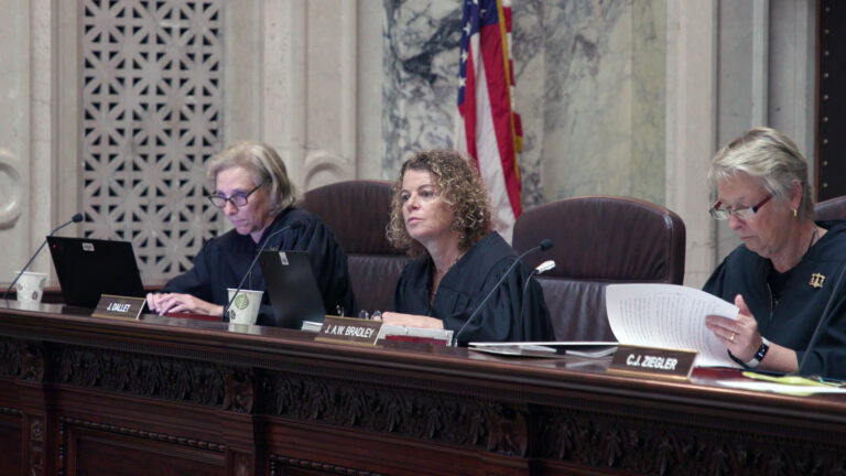 Jill Karofsky, Rebecca Dallet and Ann Walsh Bradley sit in high-backed leather chairs at a judicial bench with nameplates reading J. Dallett and J.A.W. Bradley   on its surface alongside an open laptop computer, paper documents and disposable cups, with a U.S. flag in the background of a room with marble masonry.