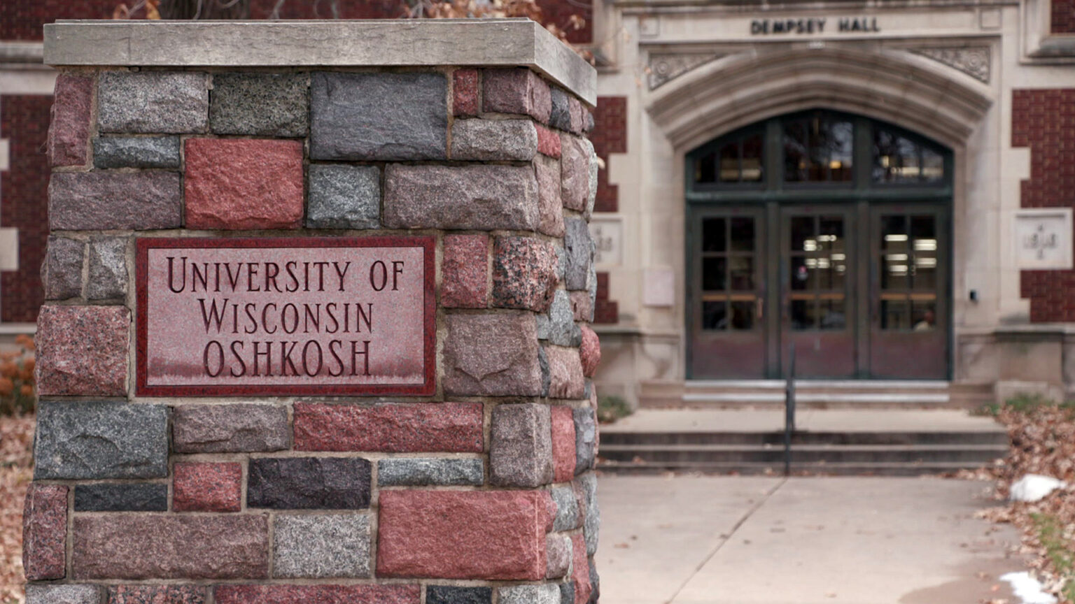 A carved stone sign reading University of Wisconsin Oshkosh is embedded among paving stones on the exterior of a pillar that stands outside a brick and masonry building with a sign above an arched entrance that reads Dempsey Hall.