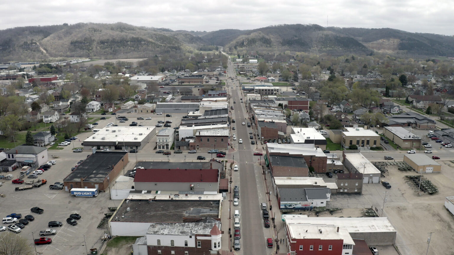 An aerial photo shows a grid of streets, buildings, parking lots and trees extending toward a series of bluffs on the horizon, under a cloudy sky.