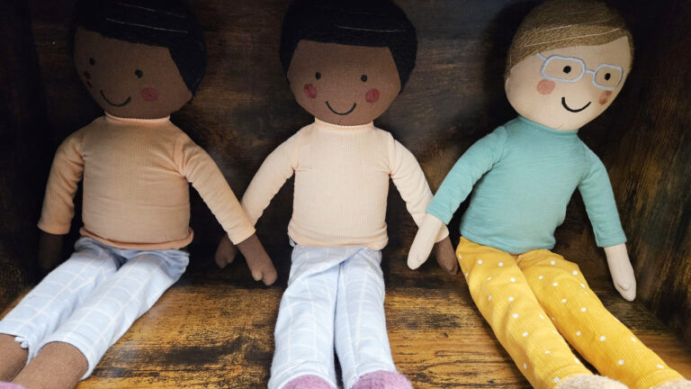 Three cloth dolls of individuals from different racial backgrounds sit on a shelf in a wood bookcase.