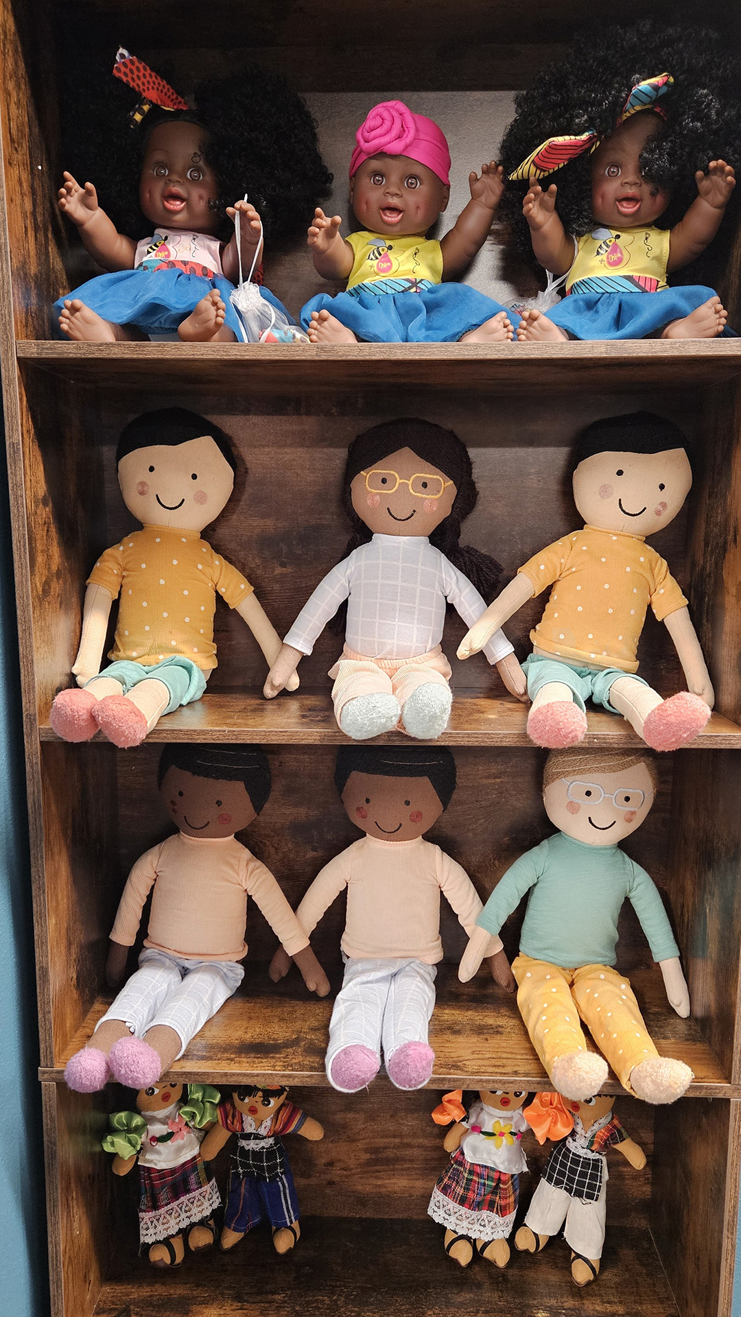 Different types of racially diverse cloth and plastic dolls sit on four shelves of a wood bookcase.