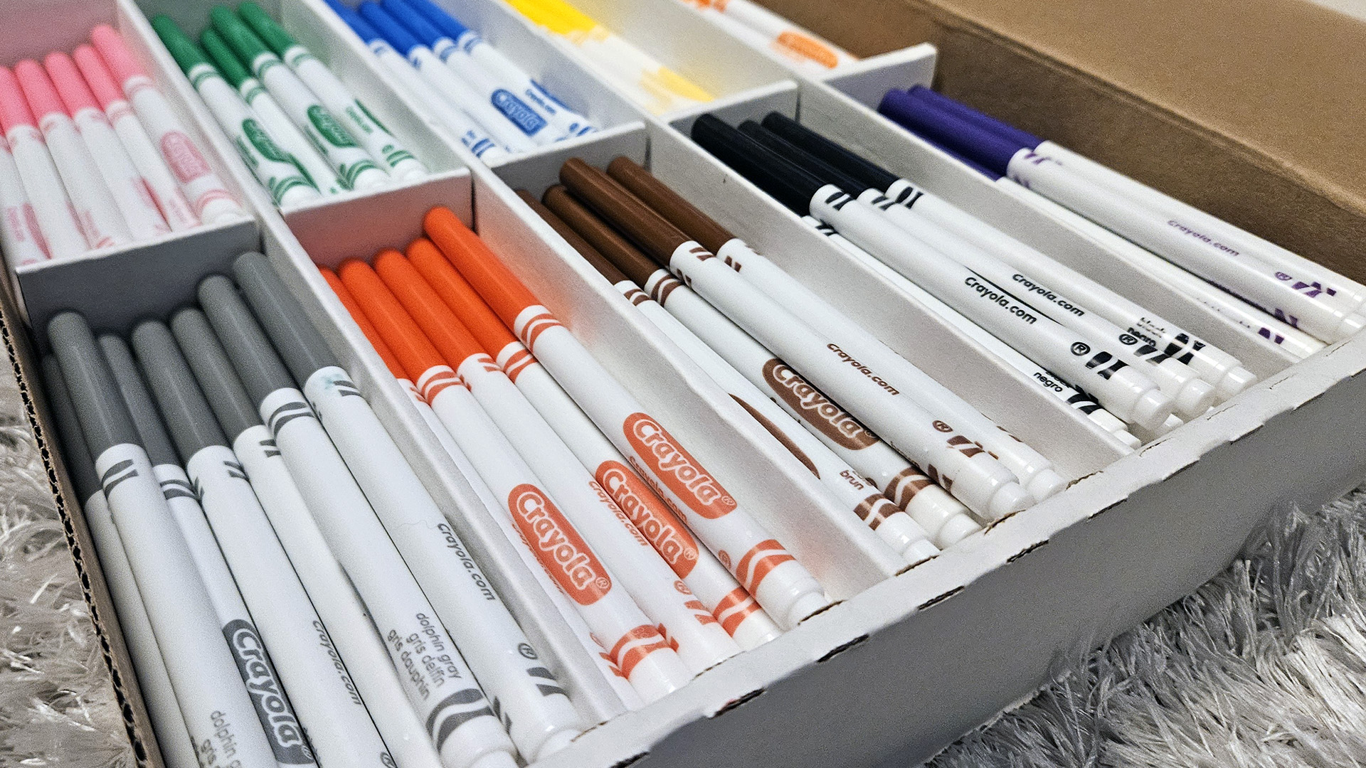 Crayola thin markers in ten different colors are organized in sections of a cardboard box sitting on top of a high pile carpet.