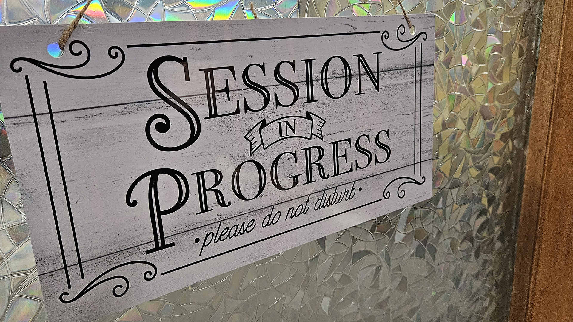 A printed plastic sign with a wood-grain laminate and the words "Session in Progress" and "please do not disturb" hangs on twine in front of a craquelure glass window in a wood door.