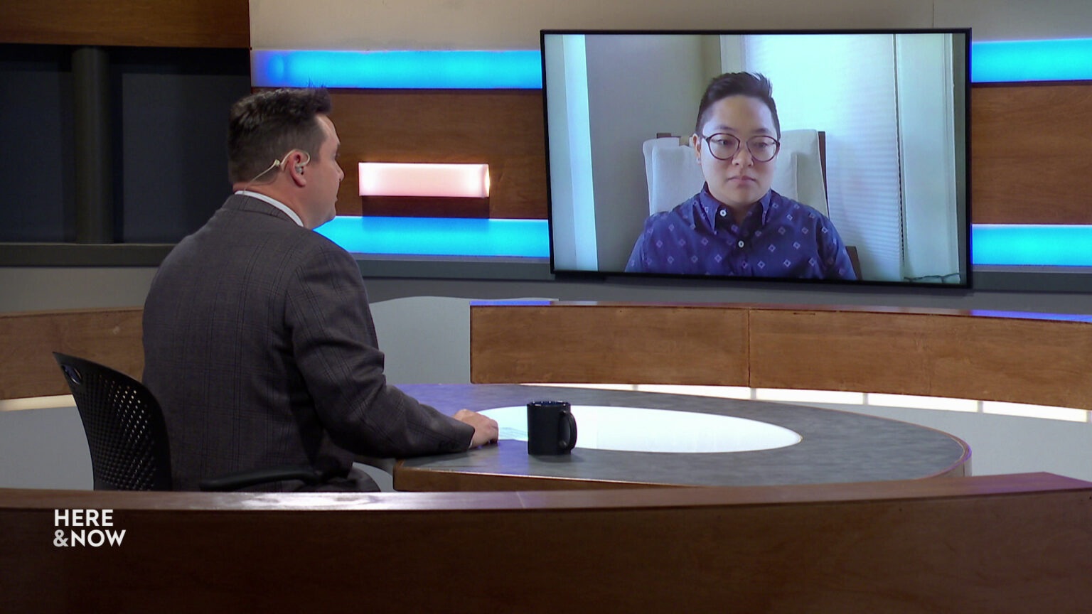 Zac Schultz sits at a desk on the Here & Now set and faces a video monitor showing an image of Reiko Ramos.