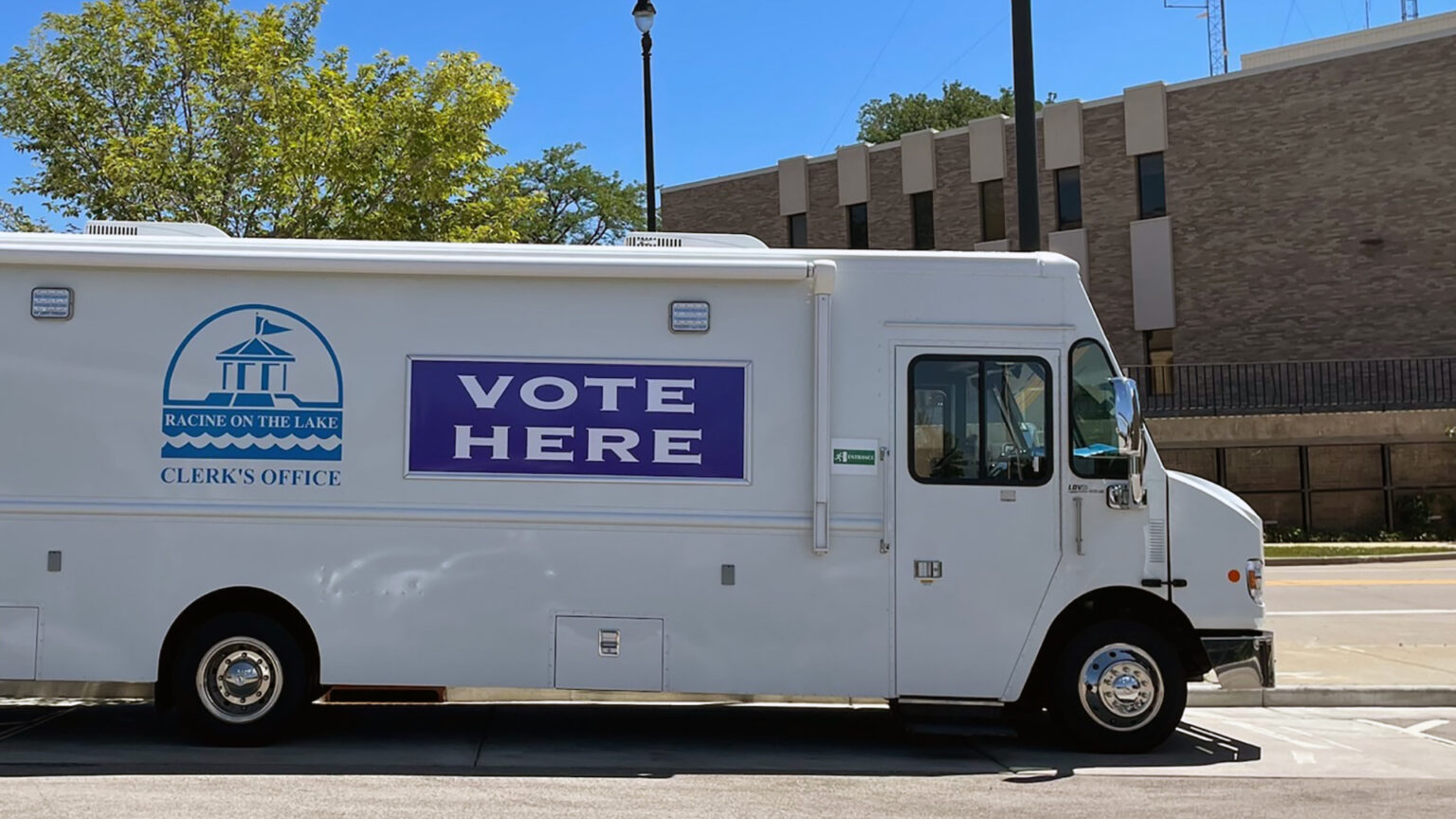 A step van with the logo of the City of Racine Clerk's office logo and the words VOTE HERE painted on its side is parked in a parking lot, with trees and a building in the background.