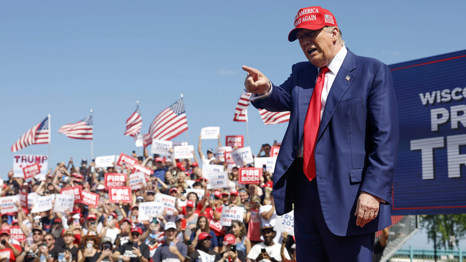 Donald Trump points with his right index finger and speaks while standing in front of seating risers filled with people holding political campaign signs, with a row of U.S. flags in the background.