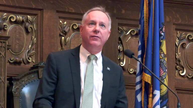 Robin Vos looks up while speaking toward a microphone, with a high-backed leather and wood chair and a Wisconsin flag behind him, in a room with wood paneling featuring carved relief crests.