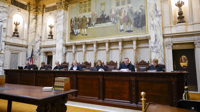 From left to right, Jill Karofsky, Rebecca Dallet, Ann Walsh Bradley Annette Ziegler, Rebecca Bradley, Brian Hagedorn and Janet Protasiewicz sit at a judicial dais in a row of high-backed leather chairs behind them, with another row of high-backed wood and leather chairs behind them, in a marble masonry room with a large wood table with law books on its surface as well as electric brass wall sconces, a U.S. flag and a large painting in the background.