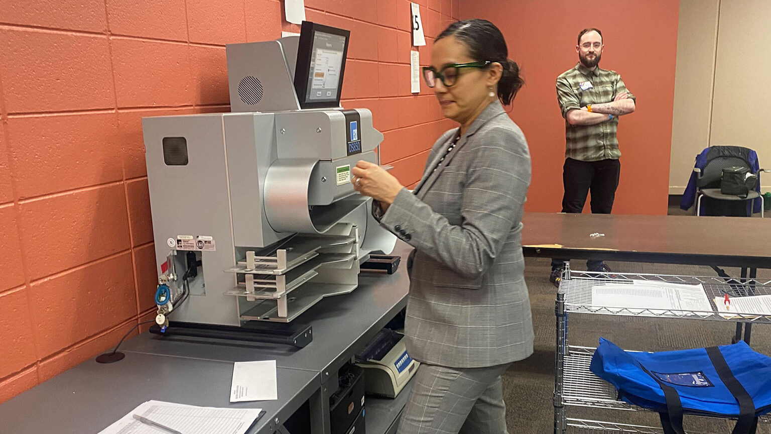 Paulina Gutierrez stands in front of a ballot tabulator machine placed on top a table, with another person standing in the background with crossed arms and wearing a disposable nametag near a chair with a jacket draped over its back, in a room with painted concrete block walls.