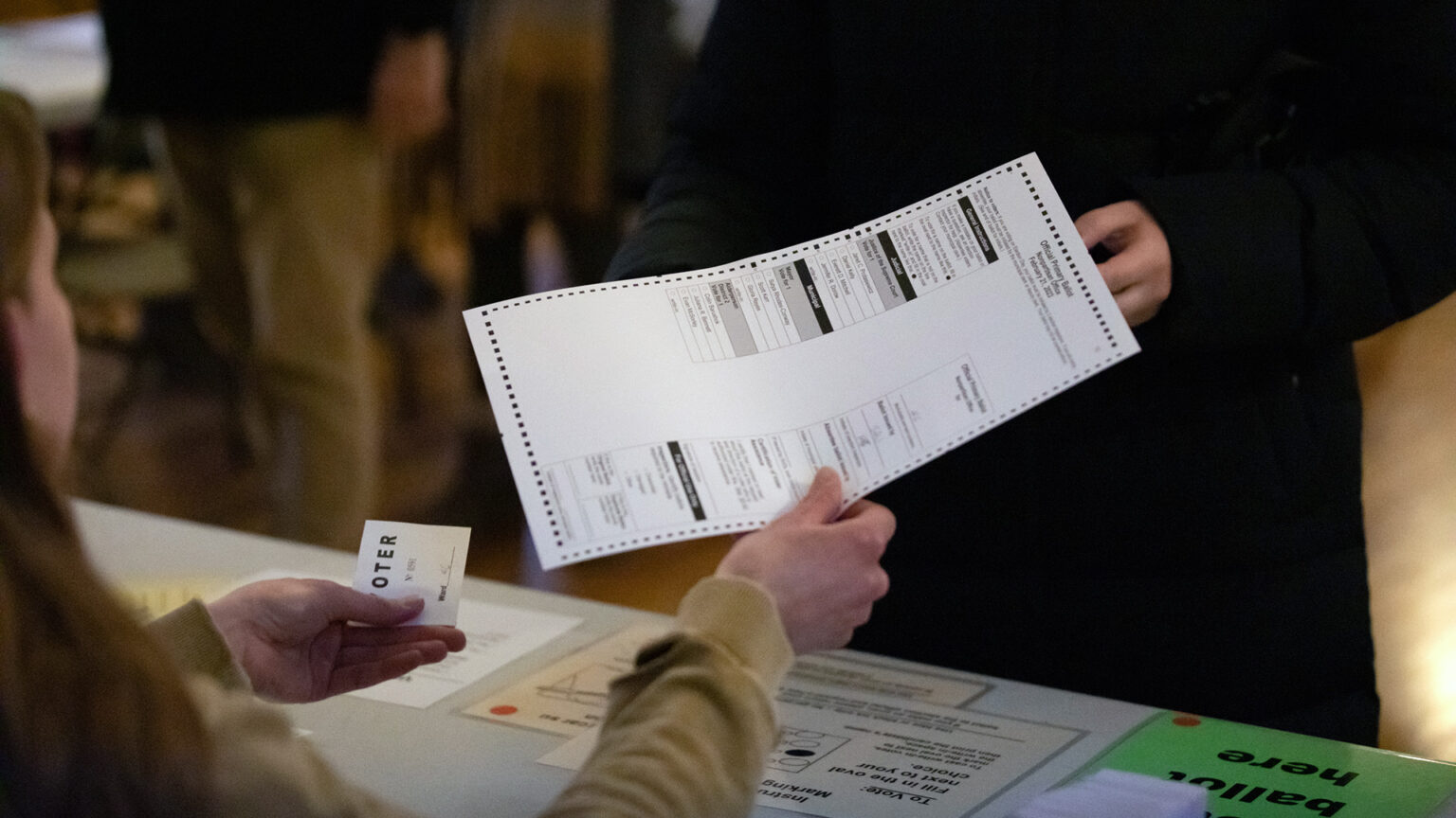 A seated person holds a voter slip in their left hand while passing a ballot in their right hand across a table to a person standing opposite them.