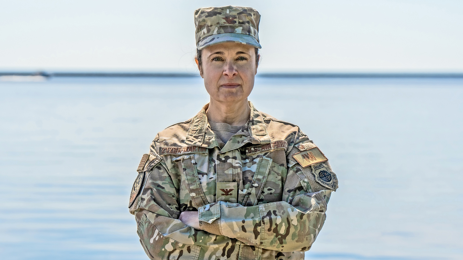 Leslie Zyzda Martin crosses her arms and poses for a portrait while wearing a combat uniform, with out-of-focus water in the background.