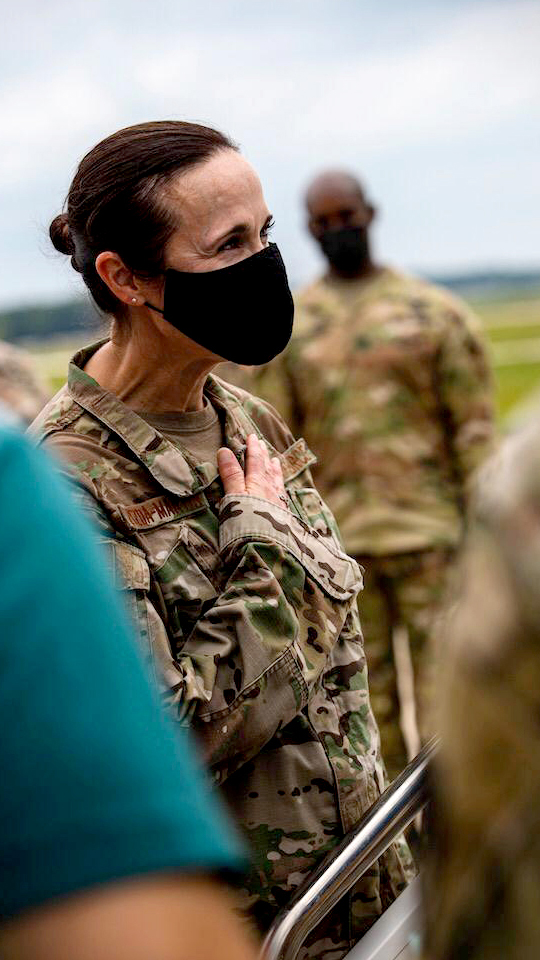 Leslie Zyzda Martin holds her right hand over her chest while wearing a medical face mask, with out-of-focus people standing in the foreground and background.