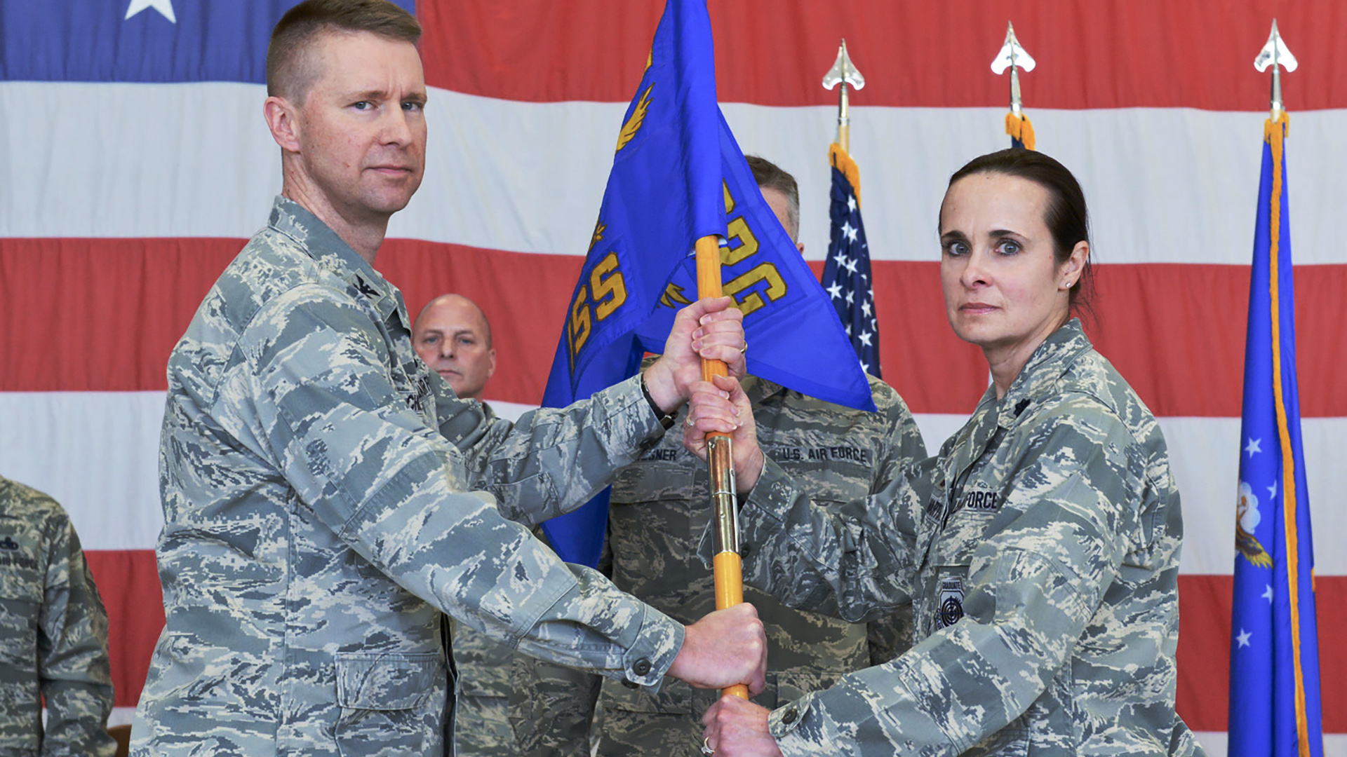 Leslie Zyzda Martin and another serviceperson wear combat uniforms while holding the pole of a unit flag, with other service people standing in the background next to multiple flags on poles and a large U.S. flag in the background.