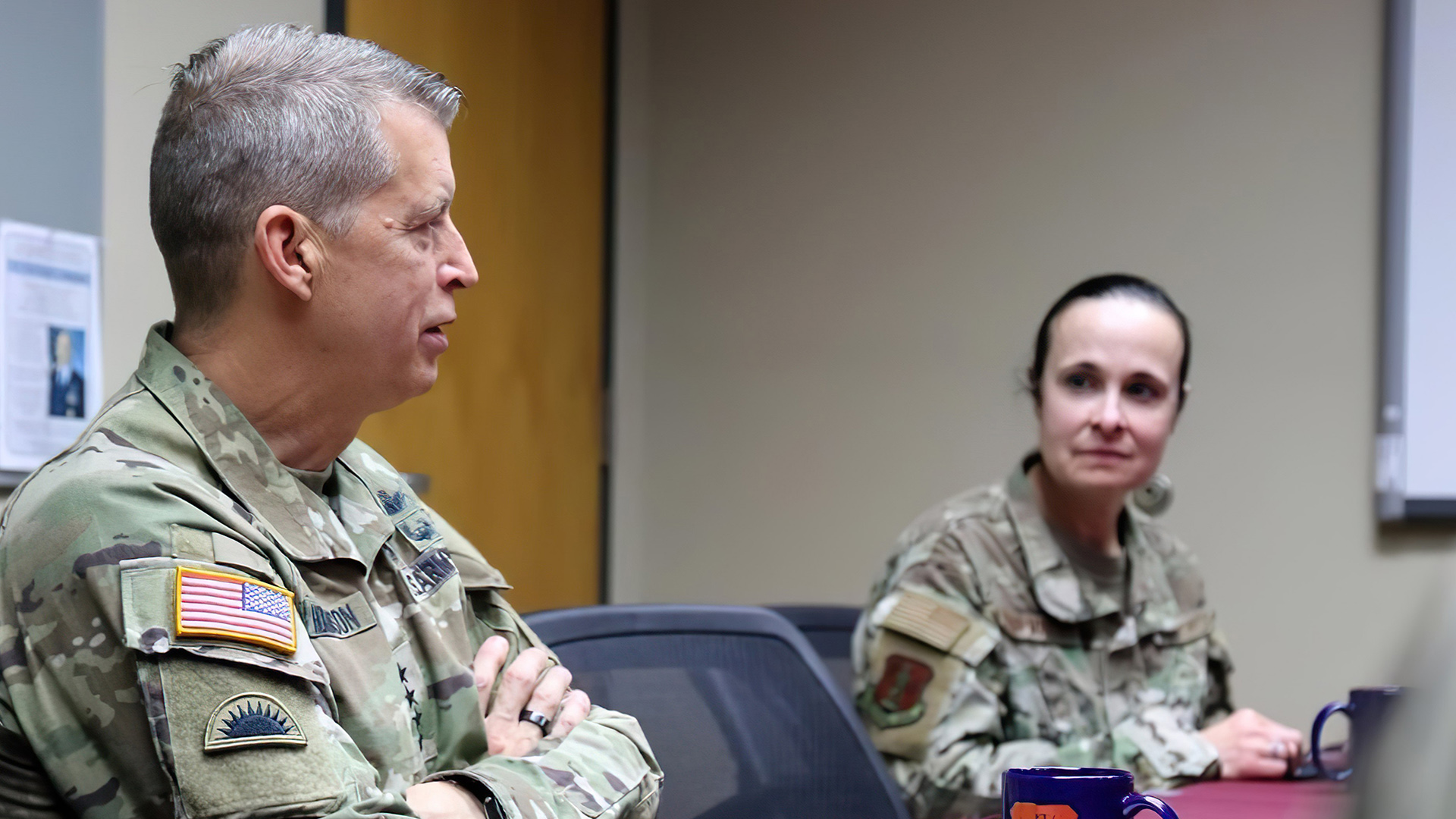 Daniel Hokanson and Leslie Zyzda Martin sit at a table with coffee mugs on its surface, in a room with a wood door, a bulletin board and a whiteboard in the background.