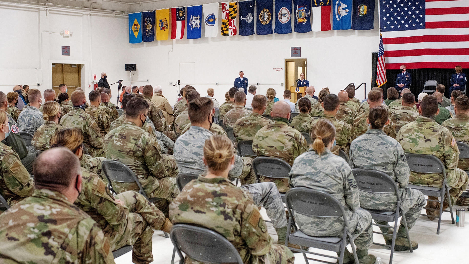 Servicepersons wearing combat uniforms sit in rows of folding metal chairs in a large room and face Leslie Zyzda Martin as she speaks at a podium while wearing a service dress uniform, with other officers wearing service dress uniforms seated on a stage with a U.S. flag on a pole and another mounted as a background, with state flags displayed in a row near the ceiling.