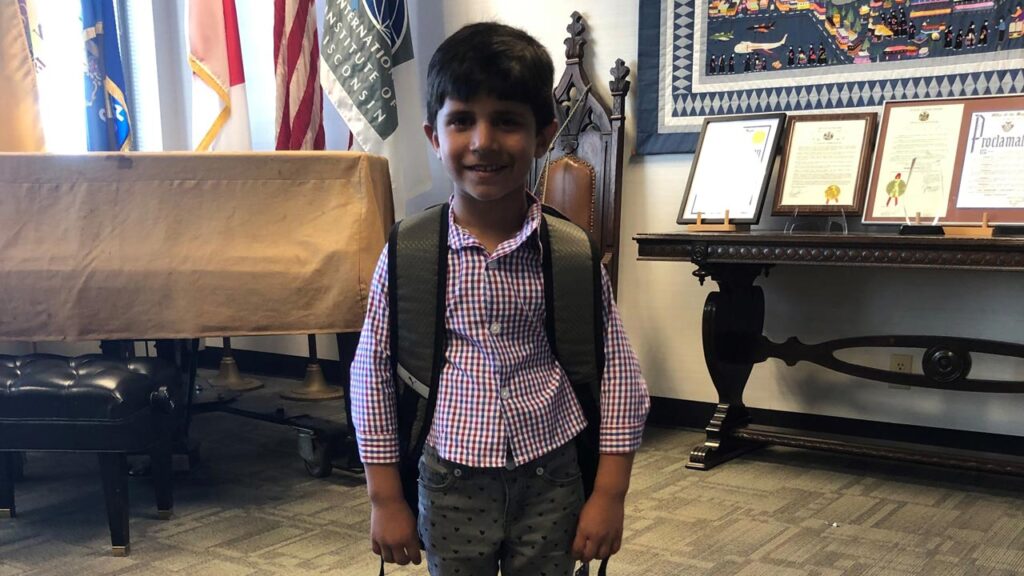 A young boy smiles, wearing a backpack in a room with flags in the background.