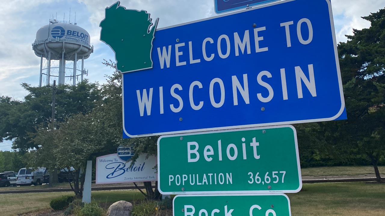 A "Welcome to Wisconsin" sign is attached to a sign that reads, "Beloit Population 36,657." In the background a Beloit water tower, trees and "Welcome to Beloit, Zonta Memorial Park" sign are in view.