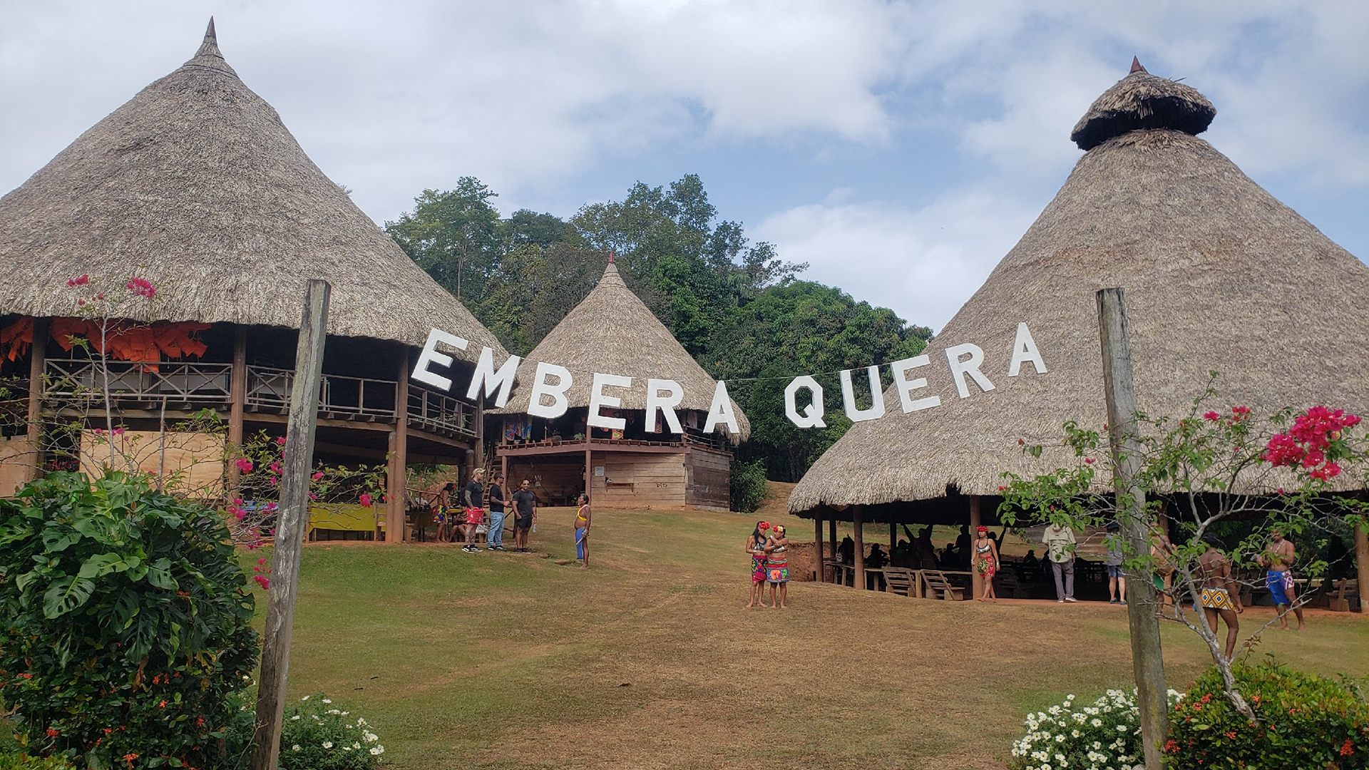Three thatched huts in a landscape in Embera Quera, Panama. Tourists and hosts stand scattered in the scene.