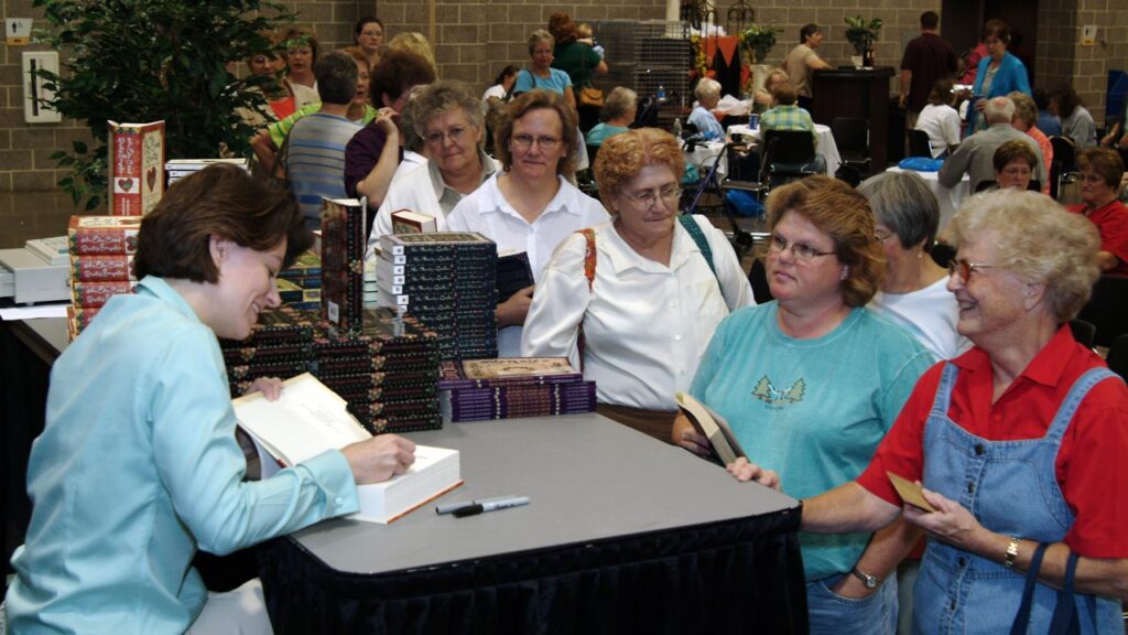 A woman signing books in a convention center, with a long line of women eager to have their books signed.