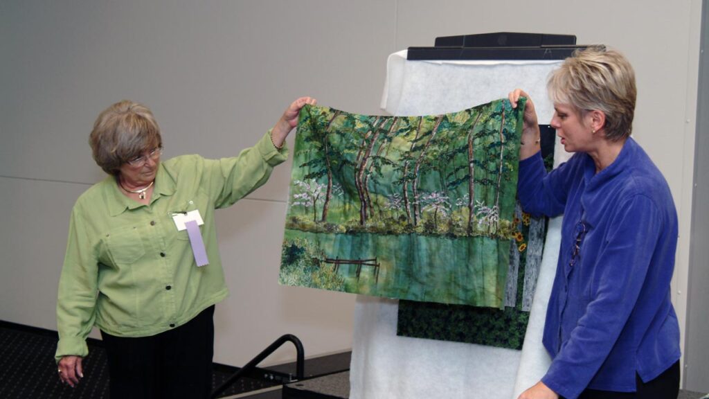 Two women hold up a lush green landscape quilt.