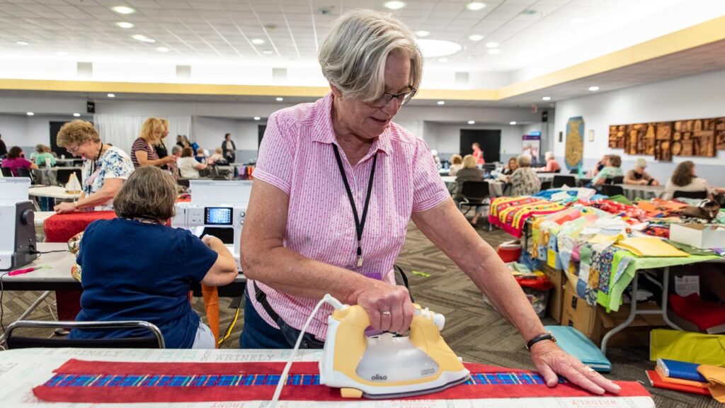 A woman irons fabric with other women at sewing machines and tables of fabric behind her.