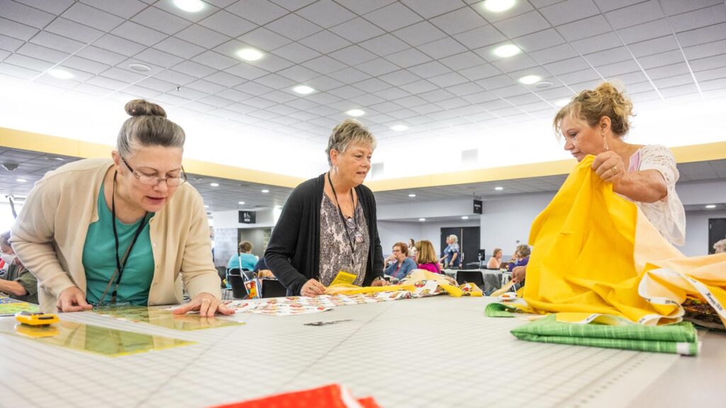 Three women cut bright fabric with quilting rulers and rotary cutters.