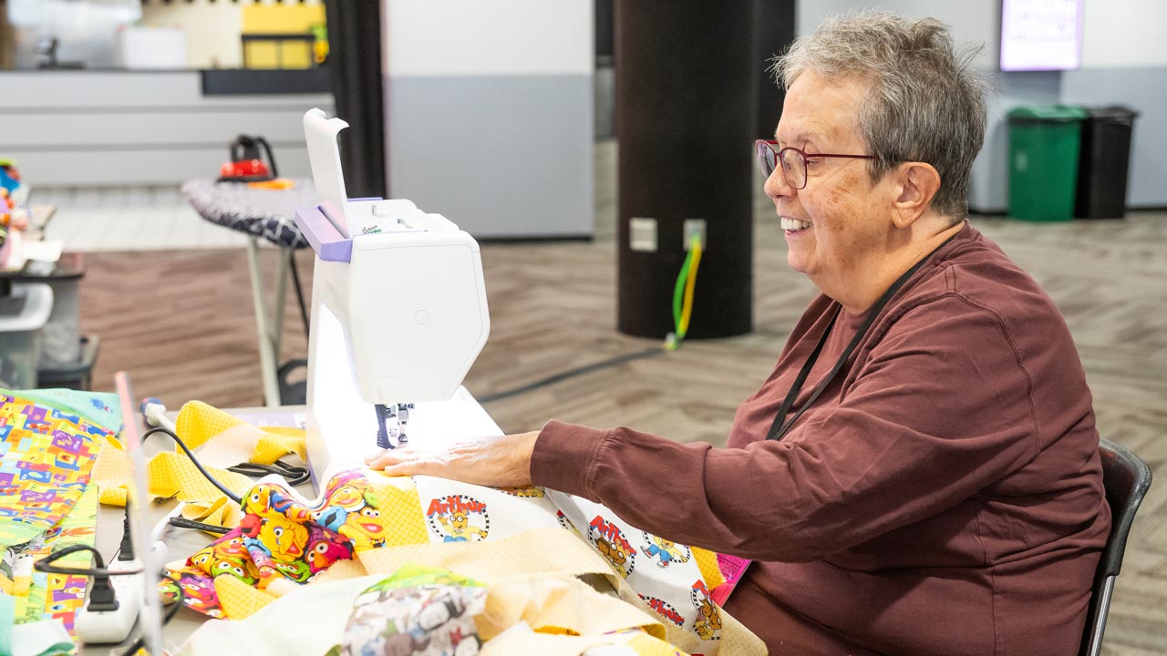 A woman smiles as she sews with PBS KIDS fabric at a sewing machine.