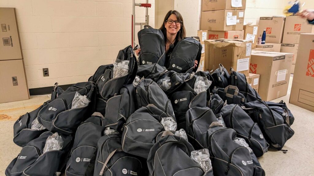 A woman kneels among a pile of backpacks with the PBS Wisconsin logo on them, the pile is up to her neck.