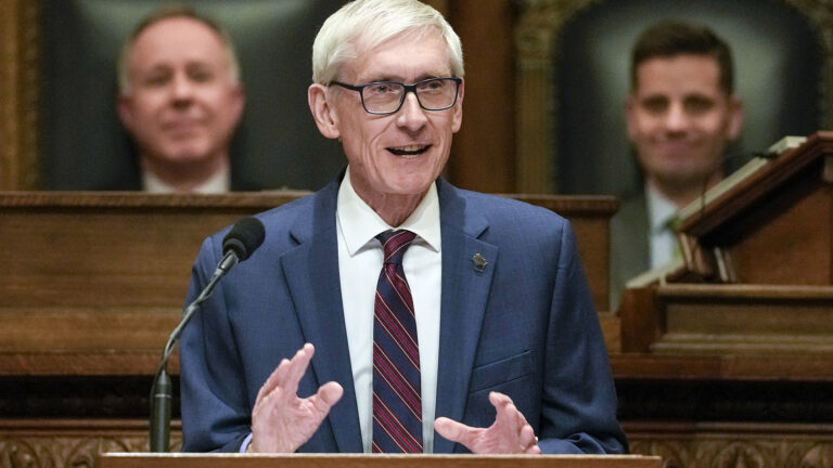 Tony Evers gestures with both hands while standing behind a podium and speaking into a microphone, with an out-of-focus Robin Vos and Chris Kapenga seated in high-backed wood and leather chairs on a higher level of a legislative dais in the background.