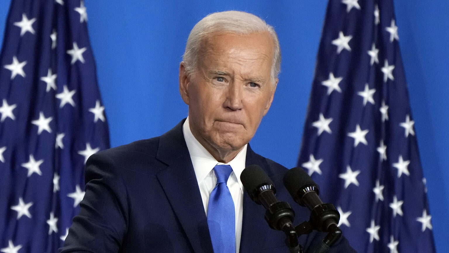 Joe Biden speaks into a pair of mounted microphones, with two U.S. flags in the background.