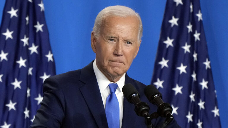 Joe Biden speaks into a pair of mounted microphones, with two U.S. flags in the background.