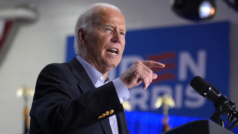 Joe Biden gestures with his right hand while standing and speaking into two microphones mounted to the top of a podium, with an out-of-focus stage light and a campaign sign in the background.