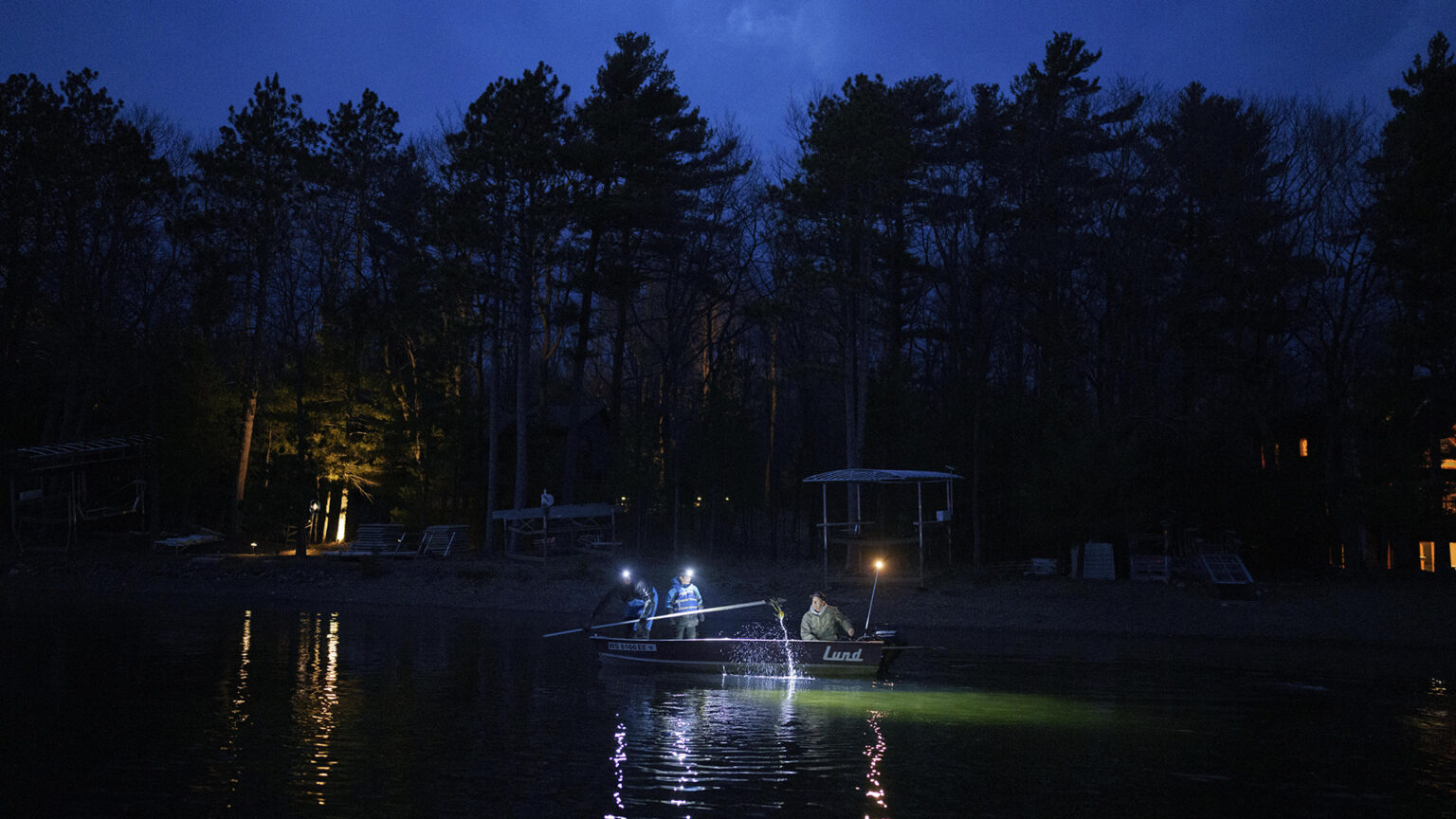 Two people wearing headlamps stand in a metal fishing boat with one spearing a fish while another person sits and steers the craft with an outboard motor on a lake in low light, with dock equipment and coniferous trees silhouetted against a cloudy, dark sky in the background.