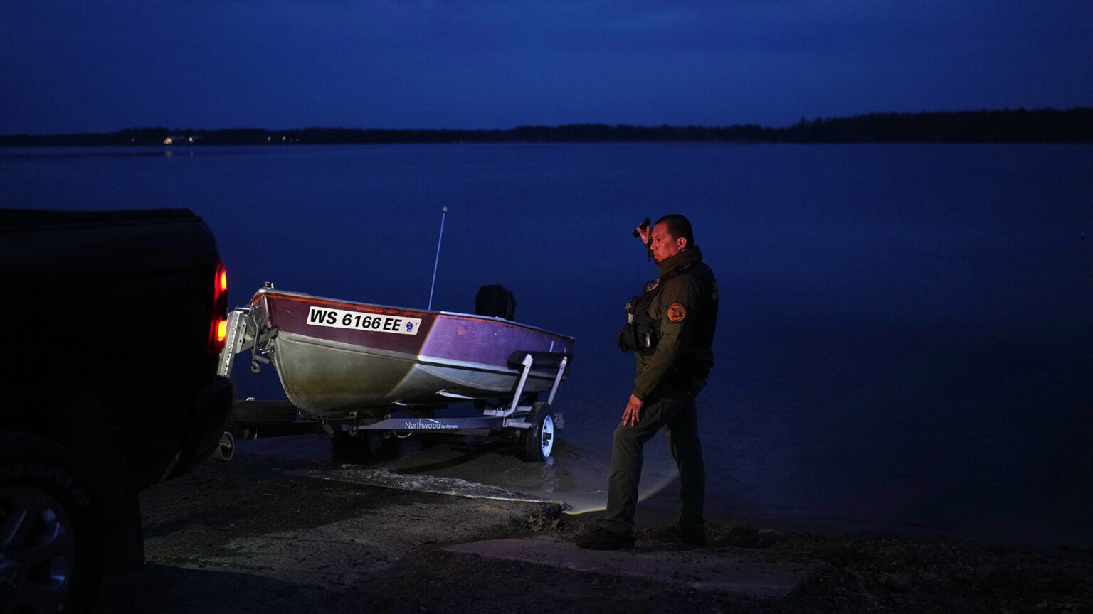 Henry Bearheart stands at the edge of a launch while a truck with illuminated rear and brake lights lowers a metal boat on a trailer into the water of a calm lake in low light, with several lights visible among numerous trees on the far shore under a cloudy, dark sky.