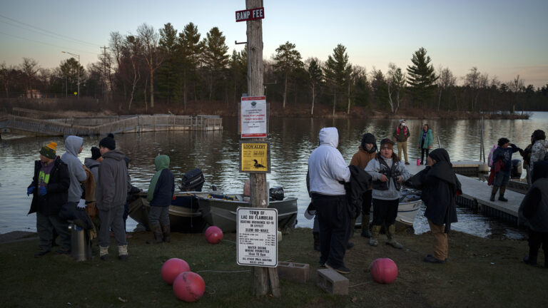 Young people stand on a floating dock and on the shore of a lake among beached metal boats with outboard motors, with several concrete block anchors connected to buoys on the ground around a wood pole with multiple signs affixed to it, and with a wooden dock railing, electric lines and trees on the opposite shore.